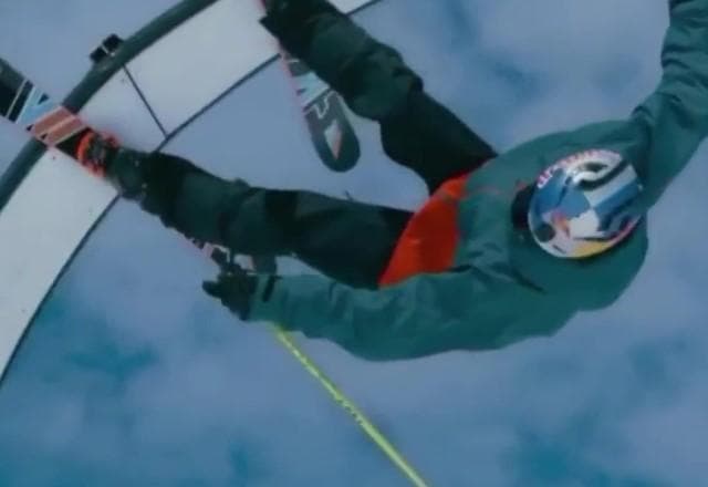 Atleta com roupa de neve, de capacete, de cabeça para baixo, fazendo manobra em uma roda e segurando um bastão, em cima de um skate de ski